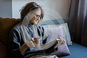 Young smiling cheerful woman in a warm sweater and eyeglasses  reading a book while sitting on the sofa with a mug of coffee