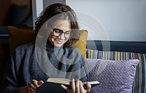 Young smiling cheerful woman in a warm sweater reading a book while sitting on the sofa