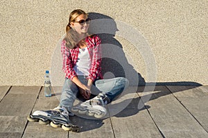 Young smiling cool girl shod in rollerblades, sits on the sidewalk and holding a water bottle.