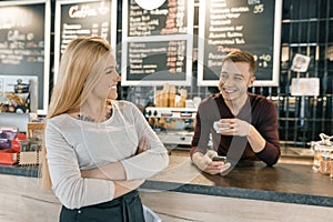 Young smiling couple of coffee shop workers, man and woman posing near the bar counter and coffee machine with cup of fresh coffee