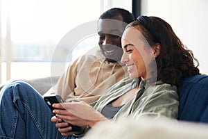 Young Smiling Couple Relaxing At Home Sitting On Sofa Checking Social Media On Mobile Phones Together