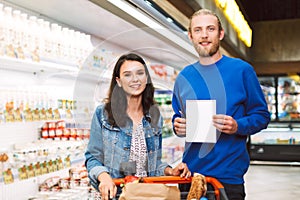 Young smiling couple with shopping list and trolley happily looking in camera in dairy department of supermarket