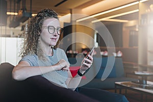 Young smiling curly business woman hipster in glasses sits on sofa in hotel lobby, cafe and holds smartphone. Girl