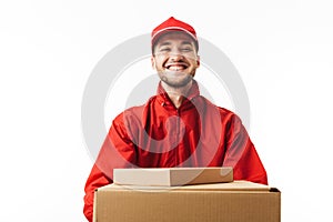 Young smiling delivery man in red cap and jacket holding boxes in hands joyfully looking in camera over white background