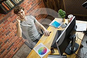 Young smiling digital artist designer talking by smartphone with his customer at his workplace in loft style office
