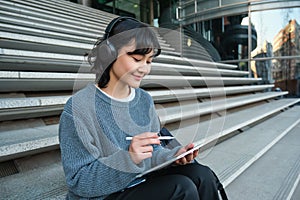 Young smiling digital artists works on her project using tablet and graphic pen, listens music in headphones and sits on