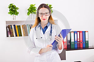 Young smiling female doctor in uniform in doctor`s ofice with clipboard