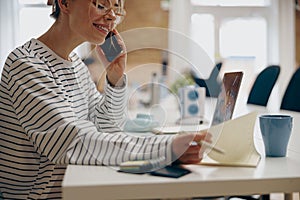 Young smiling female entrepreneur making notes while sitting in office and talking by phone