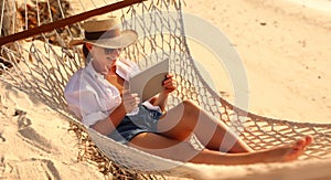 Young smiling female, happy woman in straw hat and sunglasses using digital tablet while relaxing in the hammock on tropical sandy