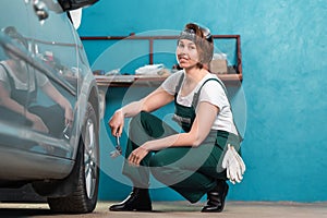 Young smiling female mechanic in blue coveralls squatting near tire and holding ratchet wrench. The concept of