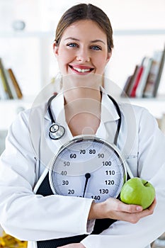 Young smiling female nutritionist holding weight scale and apple in the consultation.