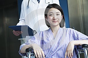 Young smiling female patient sitting in a wheelchair, doctor standing behind her, looking at camera