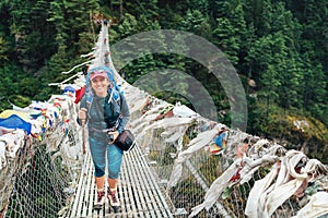 Young smiling female photographer crossing canyon over Suspension Bridge decorated with multicolored Tibetan Prayer flags hinged