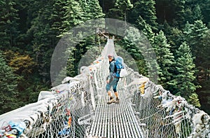 Young smiling female photographer crossing canyon over Suspension Bridge decorated with multicolored Tibetan Prayer flags hinged