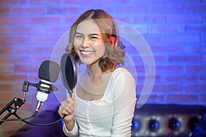 Young smiling female singer wearing headphones with a microphone while recording song in a music studio with colorful lights