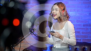 Young smiling female singer wearing headphones with a microphone while recording song in a music studio with colorful lights