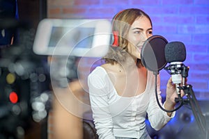 Young smiling female singer wearing headphones with a microphone while recording song in a music studio with colorful lights