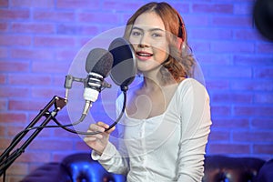 Young smiling female singer wearing headphones with a microphone while recording song in a music studio with colorful lights