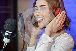 Young smiling female singer wearing headphones with a microphone while recording song in a music studio with colorful lights