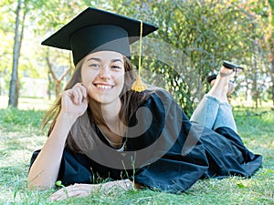 Young smiling female student in black graduate gown lies on the grass in the university park