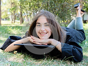 Young smiling female student in black graduate gown lies on the grass in the university park