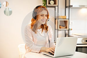 Young smiling friendly ginger girl working as agent at call center from home. Wearing headset and typing on laptop