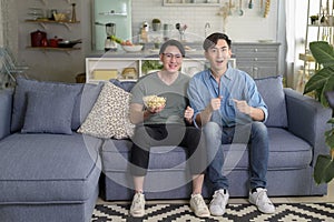 Young smiling gay couple watching TV in the living room at home, LGBTQ and diversity