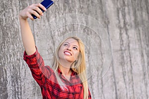 Young smiling girl with beautiful face taking self-portrait on her smartphone. She has blonde hair, beaming smile. She is wearing