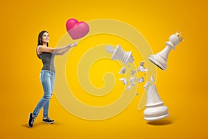 Young smiling girl in casual clothes holding big red heart, standing next to big white pawn wearing gold crown and