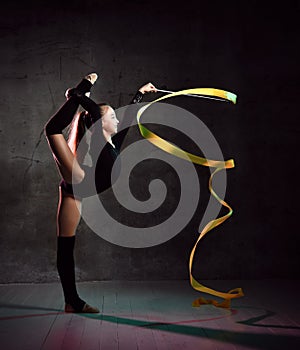 Young smiling girl gymnast in black sport body and uppers standing and making exercise with yellow gymnastic tape