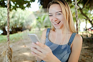 Young smiling girl riding swing and using smatrphone, sand and tree in background.