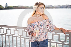 Young smiling girl sitting on the embankment of the river in the city and drinking lemonade. Outdoors lifestyle portrait