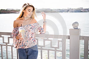 Young smiling girl sitting on the embankment of the river in the city and drinking lemonade. Outdoors lifestyle portrait