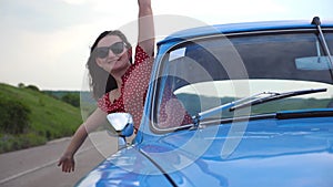 Young smiling girl in sunglasses leaning out of vintage car window and enjoying trip. Happy attractive woman looks out
