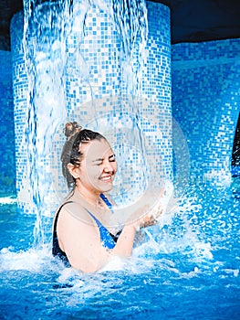 A young smiling girl swimming in the pool with blue clear water. A girl is standing under a stream of water. Pure stream