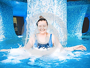 A young smiling girl swimming in the pool with blue clear water. A girl is standing under a stream of water. Pure stream