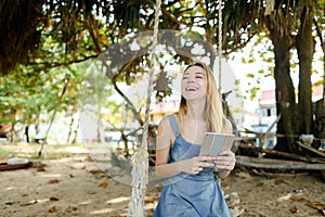 Young smiling girl using tablet and riding swing on sand, wearing jeans sundress.