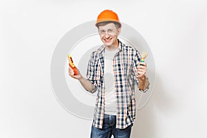 Young smiling handsome man in casual clothes and protective orange hardhat holding toy hammer and saw isolated on white