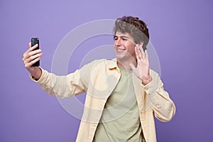 Young smiling handsome man saying hi and waving hand while making video call on smarphone isolated on purple banner background