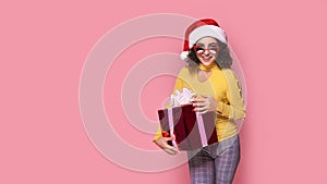 Young smiling happy cheerful curly girl in Santa's hat holds red giftbox while stands on pink background. Sopping