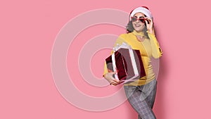 Young smiling happy cheerful curly girl in Santa's hat holds red giftbox while stands on pink background. Sopping