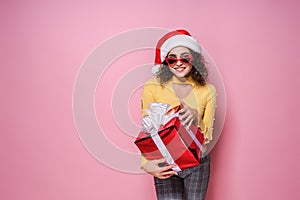 Young smiling happy cheerful curly girl in Santa& x27;s hat holds red giftbox while stands on pink background. Sopping