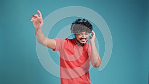 Young smiling happy cheerful student Indian man 20s dancing with headphones listen to music have fun in coral t-shirt on