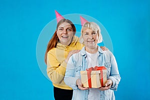 Young smiling happy daughter and mother together gifting birthday present with red ribbon isolated on blue color