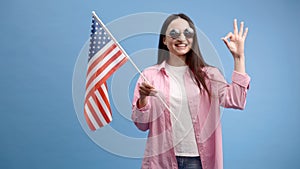 Young smiling happy woman wears casual clothes holding American flag and looking to the camera isolated on blue color background