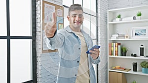 Young, smiling hispanic man confidently giving a thumb up while working on his cellphone at his office, displaying a positive,