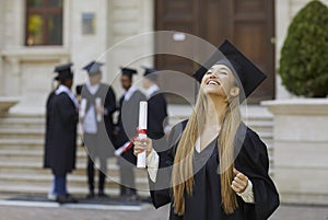 Young smiling joyful girl student in a university graduate gown and diploma in her hands.