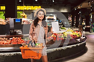 Young smiling lady in hat holding basket full of products happily looking in camera while showing shopping list in