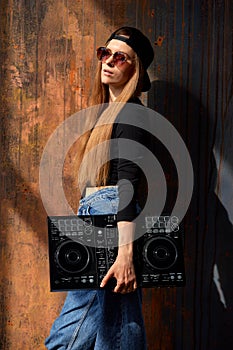 A young smiling long-haired DJ girl in a black top, jeans, a black baseball cap and sunglasses stands against a grunge