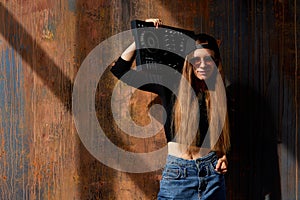 A young smiling long-haired DJ girl in a black top, jeans, a black baseball cap and sunglasses stands against a grunge